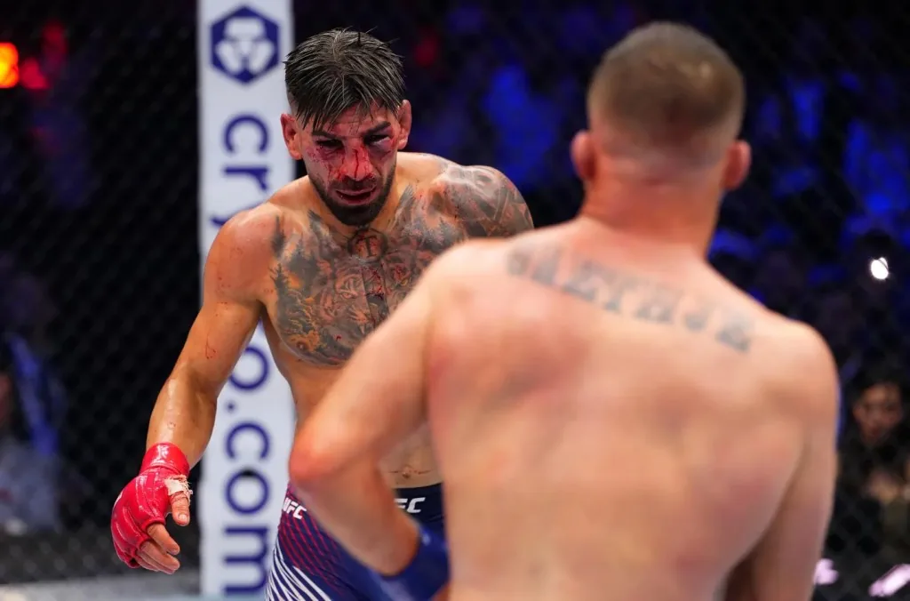 Dos luchadores de UFC se enfrentan en el octágono, uno con el rostro ensangrentado.