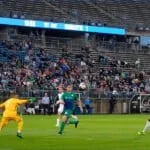 Un jugador de fútbol con uniforme amarillo disputa el balón frente a dos rivales de verde en un estadio lleno de público.