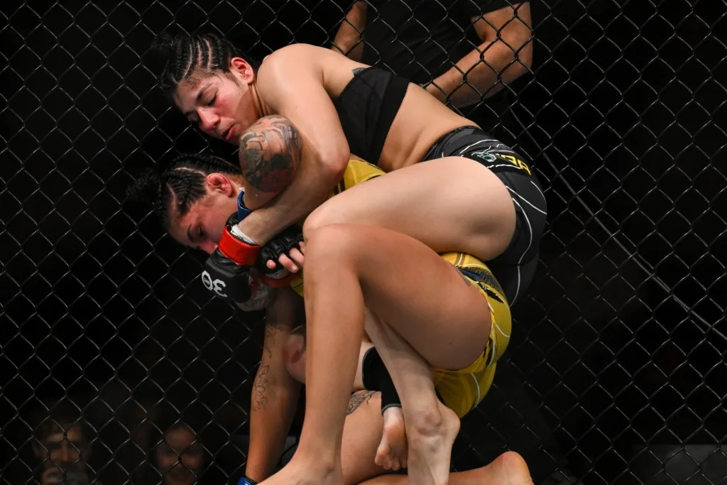 Dos luchadoras de artes marciales mixtas forcejean contra la reja del octágono durante un combate.