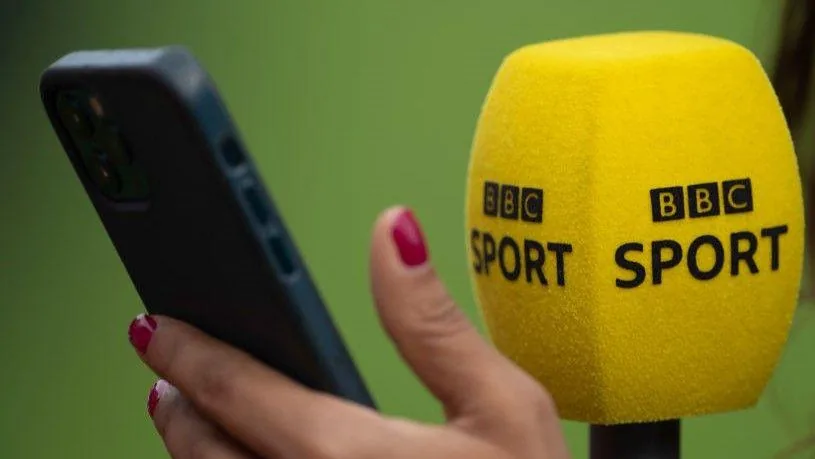 Un micrófono amarillo de BBC Sport junto a un teléfono móvil sostenido por una mano.