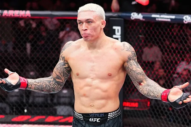 Un luchador de UFC, con el torso desnudo y los brazos tatuados, hace un gesto de duda dentro de la jaula.